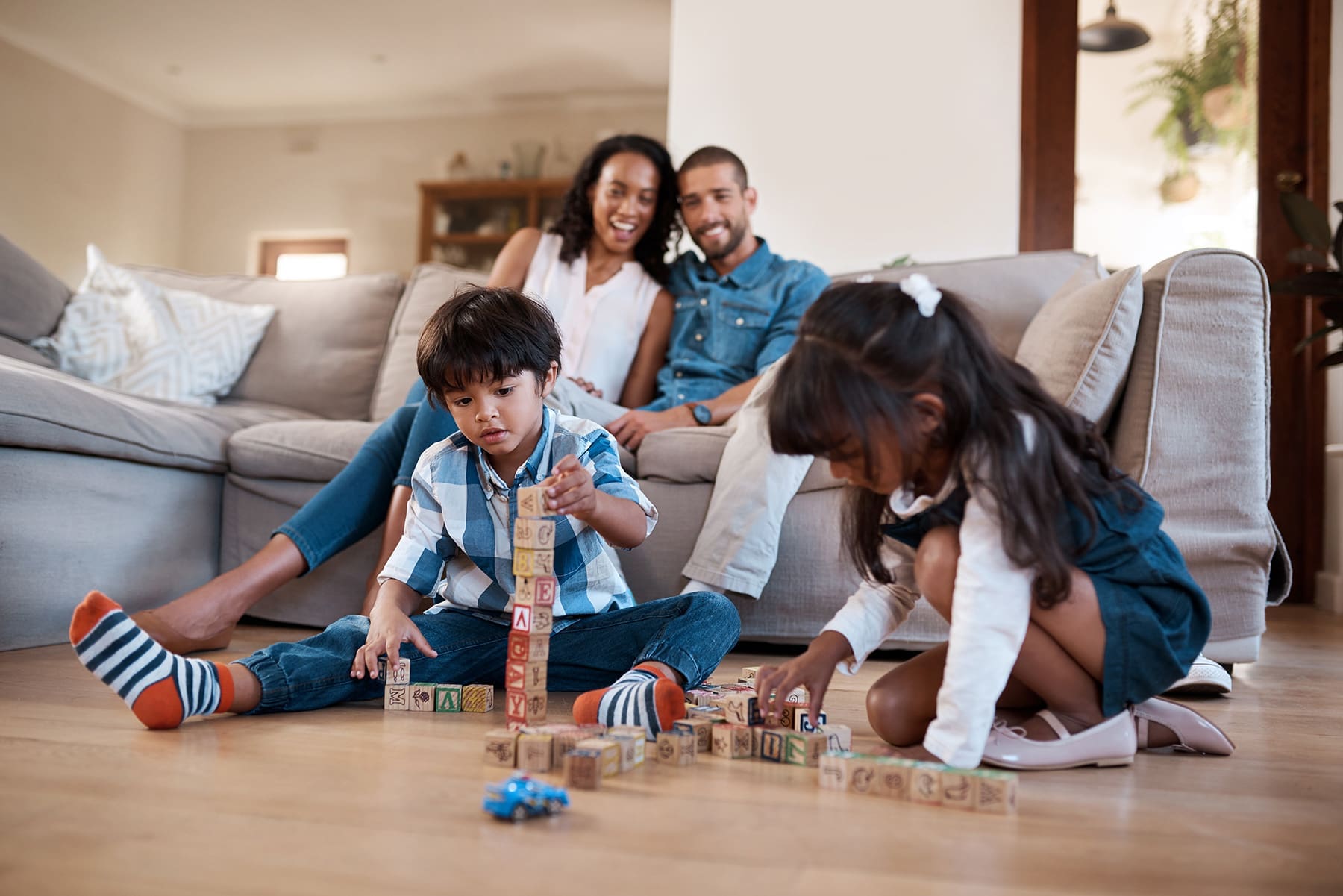 Young children playing with educational blocks on the floor while parents watch from the couch, illustrating playful learning and positive family bonding.