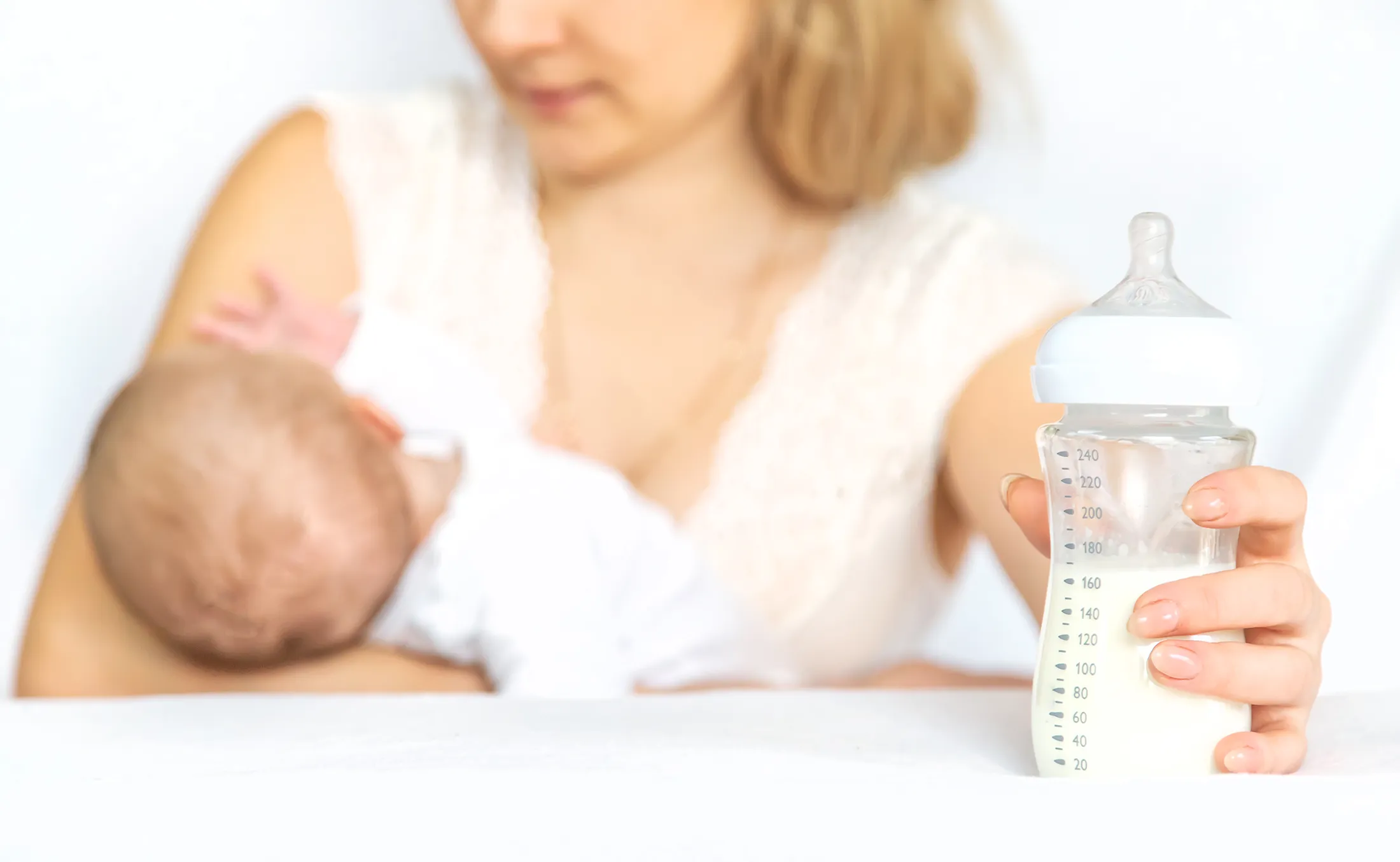 Mother breastfeeding her baby while holding a baby bottle filled with milk.