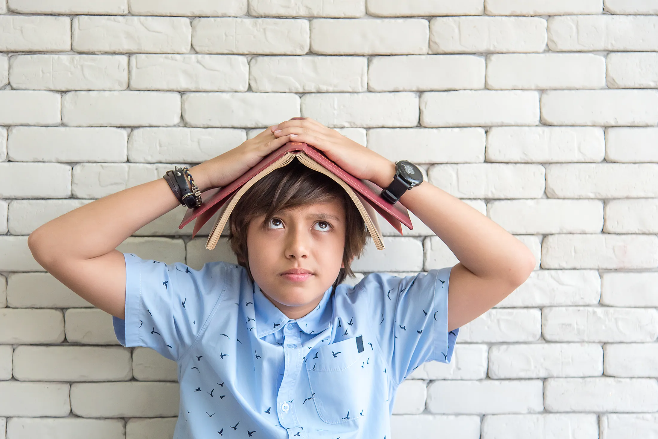 Stressed school-aged boy holding a book on his head, symbolizing the importance of stress awareness day and learning deep breathing to handle a stressful situation.