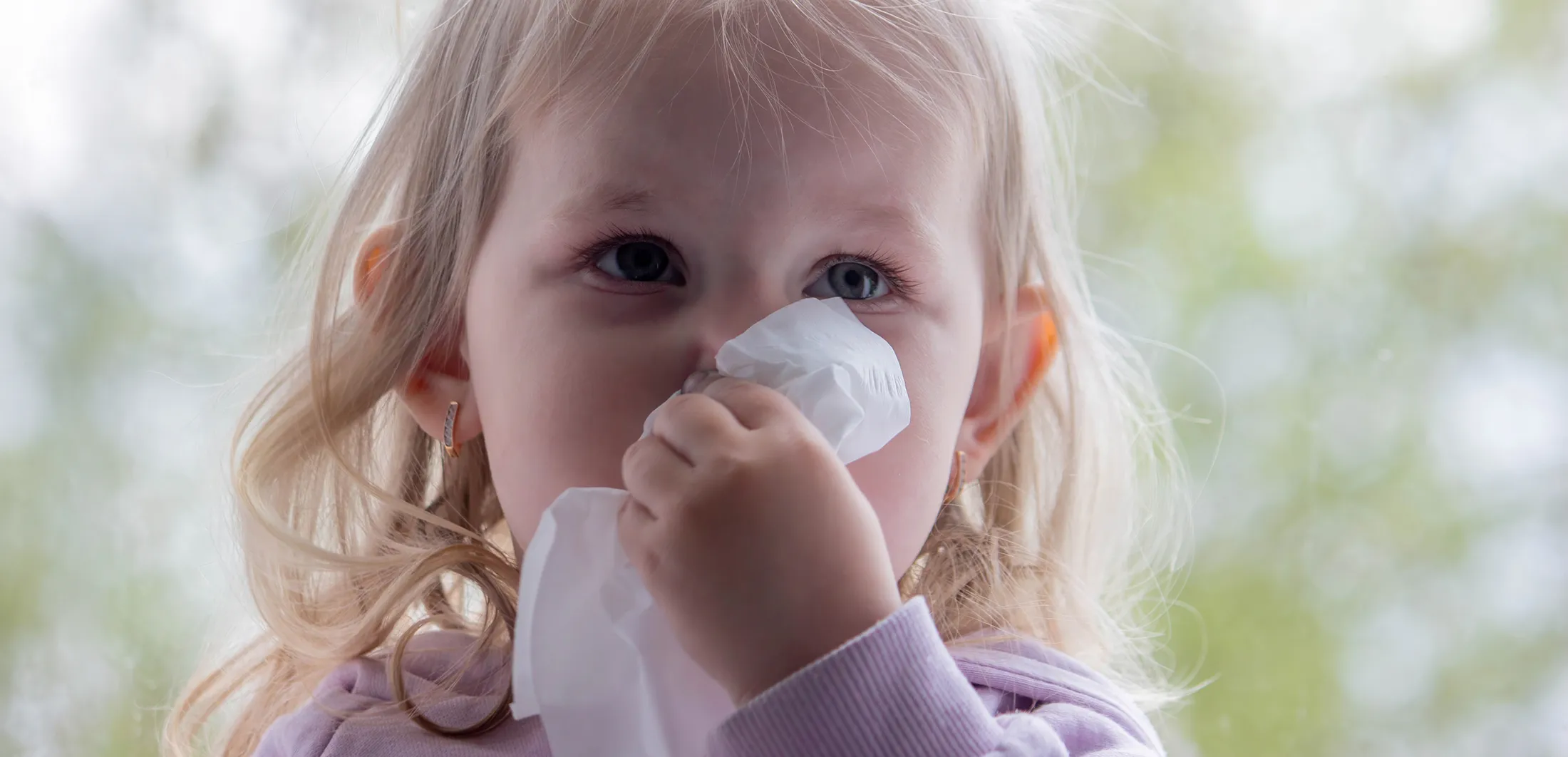 Toddler holding a tissue to their nose, showing nasal congestion commonly caused by a cold or seasonal illness.
