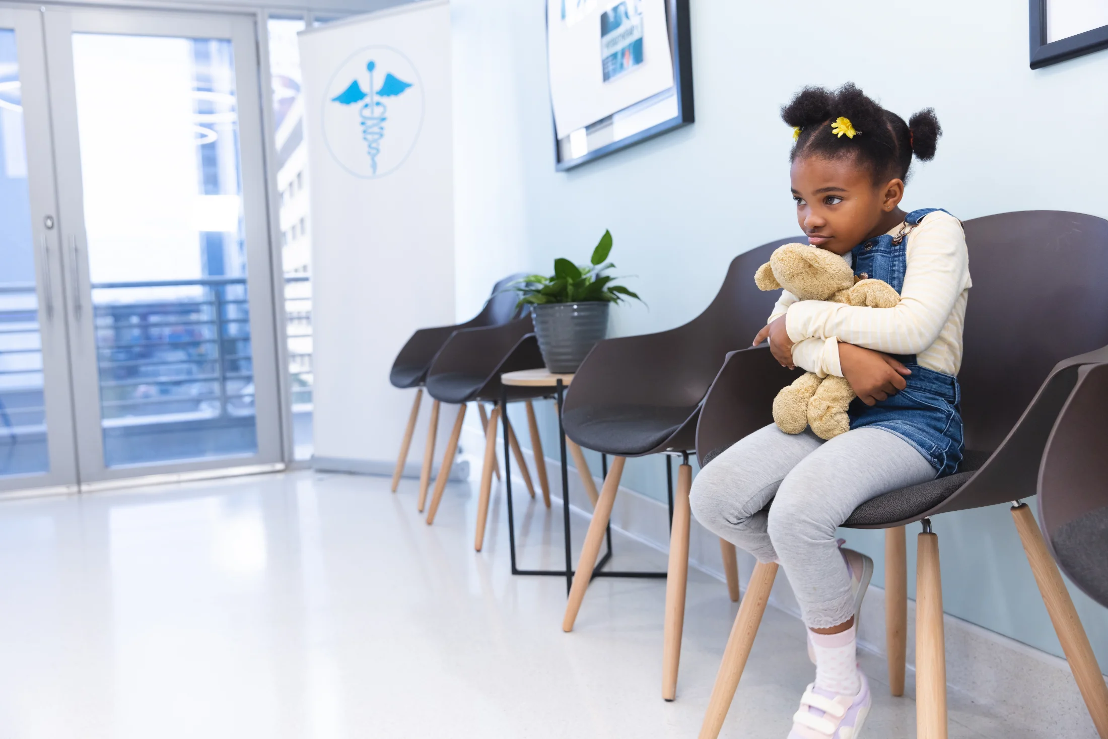 Young girl sitting in a pediatric clinic waiting room holding a teddy bear while waiting for her doctor appointment.