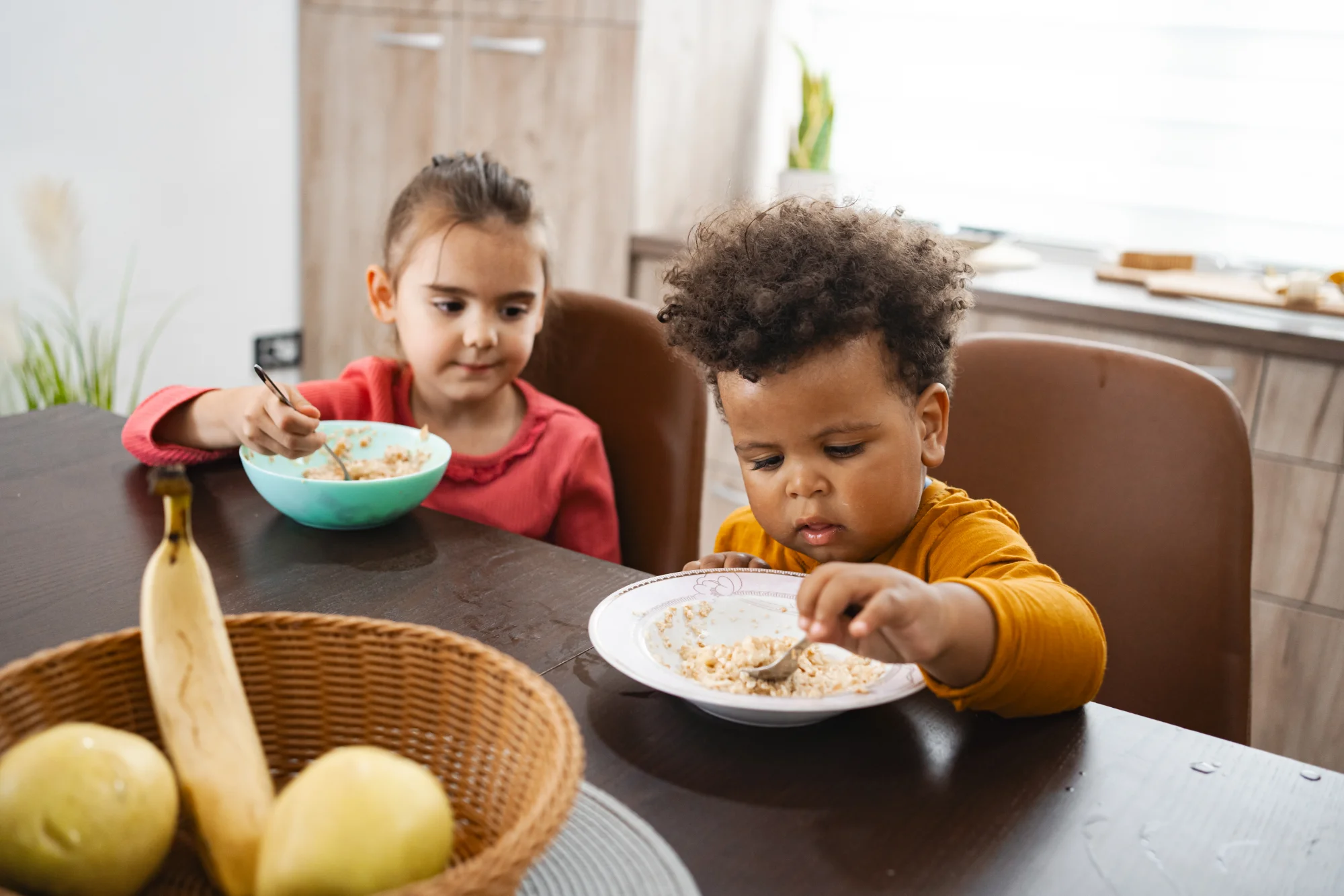 Two young children sitting at a table eating cereal, with a bowl and fruit basket in the foreground.