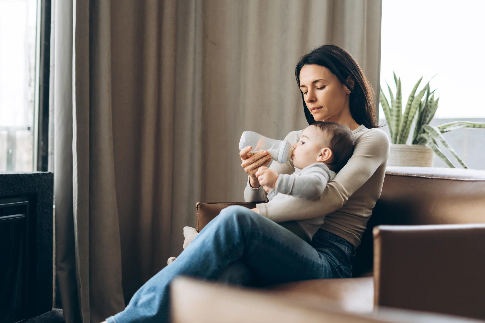 Mother sitting on a couch bottle-feeding her infant in a softly lit living room near a window