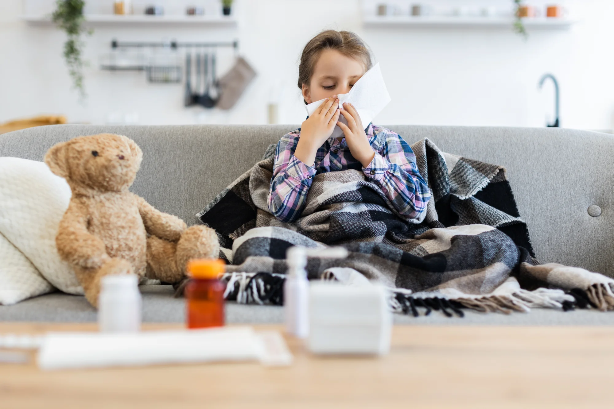 Sick young girl wrapped in blanket blowing her nose on the couch with a teddy bear and medicine nearby