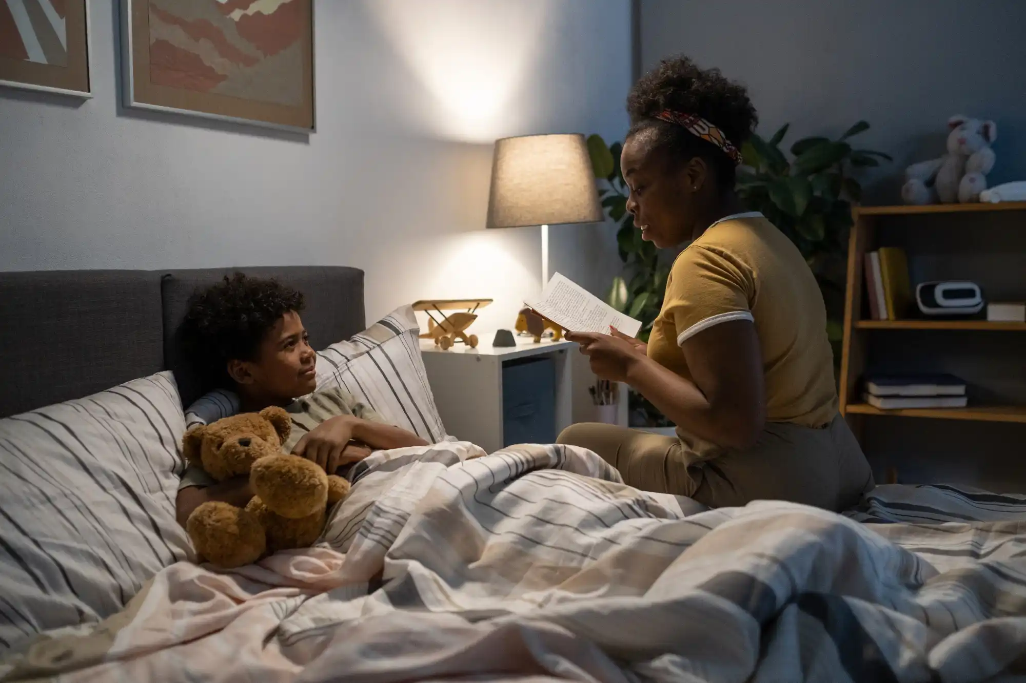Woman reading a bedtime story to a curly-haired boy tucked in bed with a teddy bear in a cozy bedroom